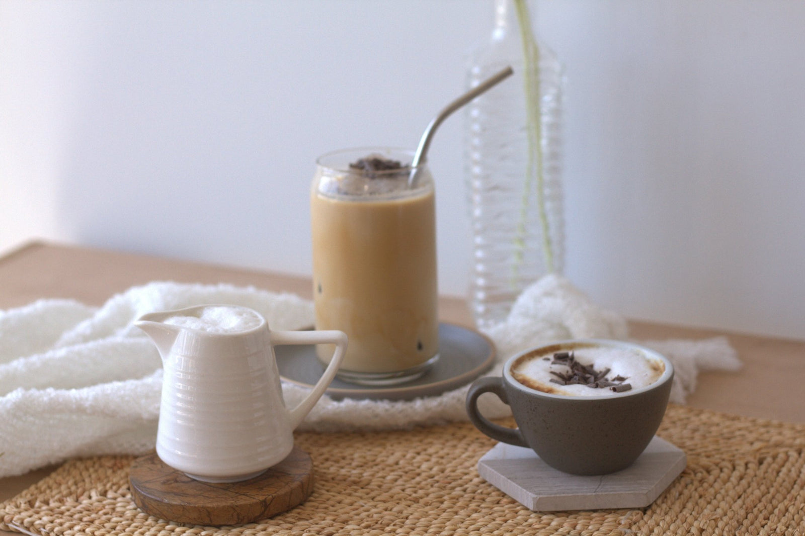 A glass of iced coffee, a mug of hot cappuccino, and a small pitcher of milk sit on a table